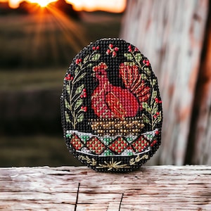 May include: An oval cross-stitch design on a black background, featuring a red rooster. The rooster is framed by a green and red floral border. The base has a gold beaded trim and a geometric pattern in red, green, and gold. It rests on a wooden surface.