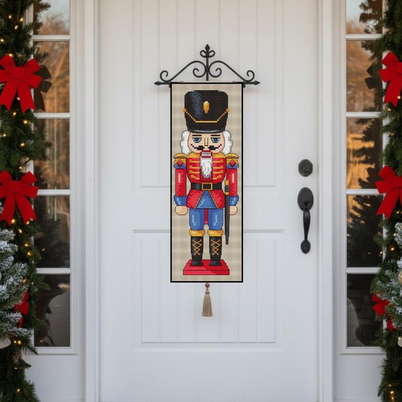 May include: A decorative Christmas door hanging featuring a colorful nutcracker soldier. The nutcracker is dressed in a red jacket, blue pants, and a black hat. The hanging is framed in black and has a tassel at the bottom.