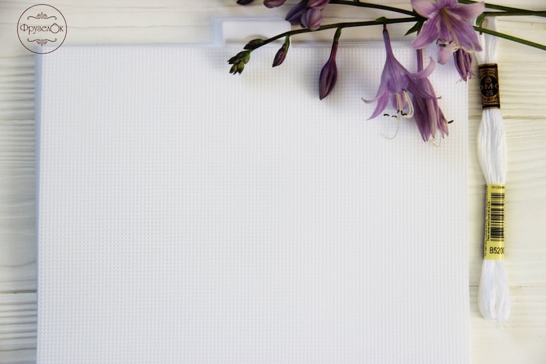 Może przedstawiać: Biała tkanina Aida z fioletowym kwiatem i białym motkiem nici do haftu na białym drewnianym tle. Moteczek nici ma ż&oacute;łtą etykietę z czarnym tekstem.