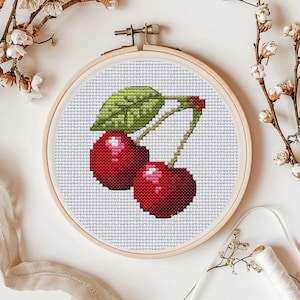 May include: Cross-stitch embroidery hoop featuring a design of two red cherries with green leaves and stems.