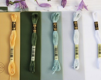 Puede incluir: Cinco madejas de hilo de bordar en diferentes colores: amarillo claro, verde, azul claro, blanco y azul claro. Cada madeja está etiquetada con un número DMC. Las madejas están dispuestas en una fila sobre una superficie blanca.