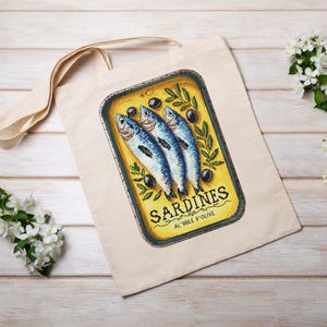 May include: A white canvas tote bag featuring a yellow and blue illustration of a tin of sardines. The tin has the text "SARDINES AL'HUILE D'OLIVE" on it.