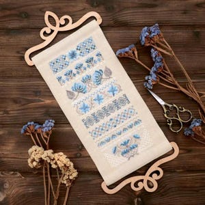 Peut inclure: Un échantillon de point de croix beige avec des motifs floraux et géométriques bleus, présenté sur un support enrouleur en bois. L'échantillon présente divers motifs brodés. Des fleurs séchées et des ciseaux décoratifs sont disposés sur une surface en bois.
