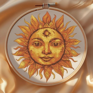 May include: A cross-stitch embroidery hoop with a sun face design. The sun has a smiling face with detailed features and is surrounded by stylized flames in shades of yellow and orange. The embroidery is set in a wooden hoop.