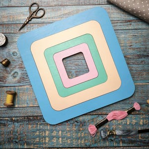 Op de afbeelding: Een gelaagde, vierkante houten knutselframe in pastelkleuren blauw, cr&egrave;me, groen en roze. Naaibenodigdheden, waaronder een schaar, garenspoelen en borduurgaren, zijn gerangschikt rond het frame op een verweerd blauw houten oppervlak.