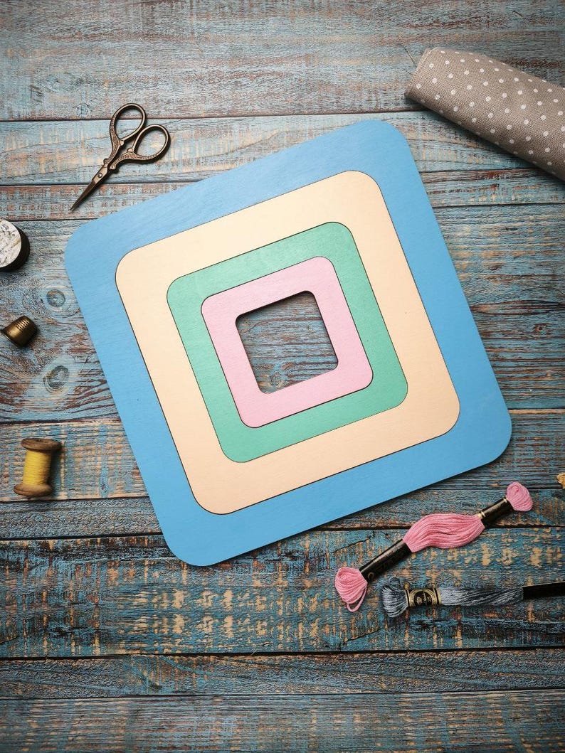 Op de afbeelding: Een gelaagde, vierkante houten knutselframe in pastelkleuren blauw, cr&egrave;me, groen en roze. Naaibenodigdheden, waaronder een schaar, garenspoelen en borduurgaren, zijn gerangschikt rond het frame op een verweerd blauw houten oppervlak.