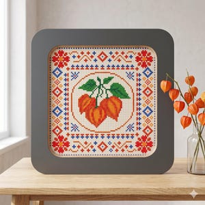 Könnte beinhalten: Kreuzstich-Kunstwerk in einem grauen Rahmen. Das Design zeigt orangefarbene und grüne botanische Elemente in einem kreisförmigen Rahmen, umgeben von einem geometrischen Muster in Rot, Blau und Orange. Das Kunstwerk wird auf einem Holztisch ausgestellt.