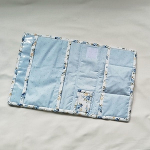 May include: A quilted organizer in light blue with white polka dots and floral accents. The edges are trimmed with a floral pattern. Features include a clear pocket, a hook and loop closure, and multiple pockets.