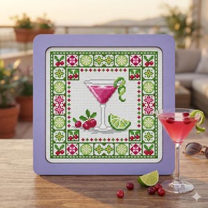 Pode incluir: Um bordado em ponto cruz quadrado numa moldura roxa clara com um cocktail cor-de-rosa num copo de martini, uma fatia de lima e bagas vermelhas. O design é rodeado por uma borda decorativa de motivos geométricos e de fruta verdes e cor-de-rosa.