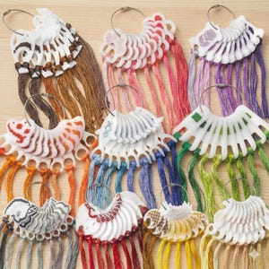 Puede incluir: Una colección de tarjetas de colores de hilo de bordar, cada una con un soporte de plástico blanco en forma de abanico y un anillo de metal. Los colores del hilo incluyen tonos de marrón, rosa, morado, naranja, azul, verde y amarillo. Las tarjetas tienen diseños bordados.