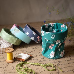 Può includere: Un cestino di tessuto verde acqua con un motivo floreale bianco e rosa. Dietro il cestino ci sono diversi anelli di tessuto con motivi floreali e a pois. Una bobina di filo giallo, una bobina di filo bianco e filo verde sono in primo piano.