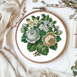 May include: A white embroidery hoop with a cross-stitch design of a bouquet of white and cream roses with green leaves.