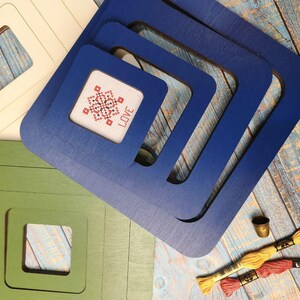 Op de afbeelding: Vierkante houten frames in cr&egrave;me, groen en blauw, met een kruissteekontwerp met de tekst "LOVE". De frames zijn genest, met borduurgaren en een vingerhoed, wat wijst op een knutselproject. Verschillende maten.