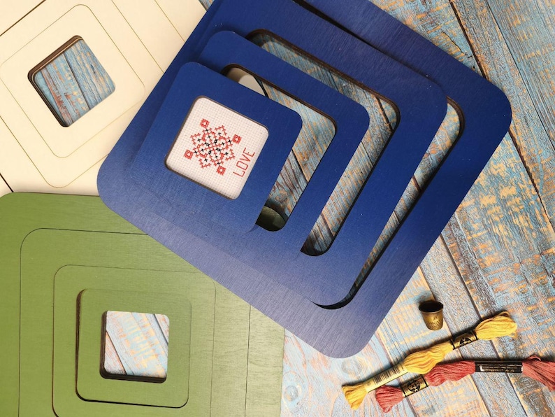 Op de afbeelding: Vierkante houten frames in cr&egrave;me, groen en blauw, met een kruissteekontwerp met de tekst "LOVE". De frames zijn genest, met borduurgaren en een vingerhoed, wat wijst op een knutselproject. Verschillende maten.