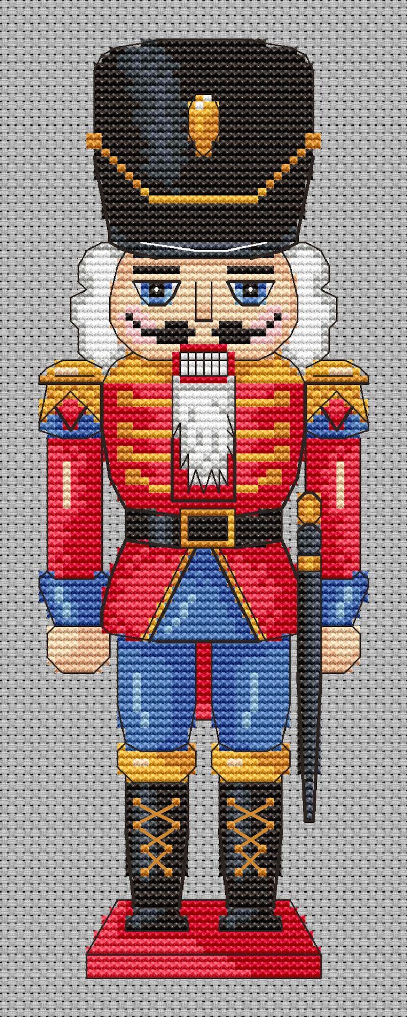 May include: A cross-stitch pattern of a traditional nutcracker soldier. The figure is dressed in a red jacket with gold accents, blue pants, and a black hat. It has a white beard and mustache, and is holding a black sword.