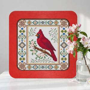 Puede incluir: Un bordado de punto de cruz enmarcado de un cardenal rojo posado en una rama con flores rosas. La obra de arte está dentro de un marco de madera rojo. El diseño de punto de cruz presenta un borde detallado con patrones geométricos y florales.