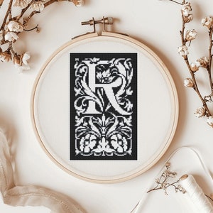 Könnte beinhalten: Schwarzer und weißer Stickrahmen mit einem Kreuzstich-Design des Buchstabens "R" in einem dekorativen floralen Rahmen.