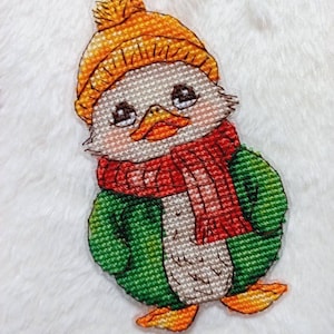 May include: A cross-stitch embroidery of a duck wearing a green jacket, a red scarf, and an orange hat with a pom-pom.