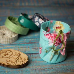 May include: A selection of handmade fabric containers and coasters. The main container is light blue with a floral pattern. Other containers feature green, white, and navy patterns. A wooden coaster with a floral design is also present.