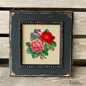 May include: A framed cross-stitch floral design featuring a bouquet of red and pink roses, with purple flowers and green leaves. The artwork is set against a cream background and framed in a distressed black wooden frame.