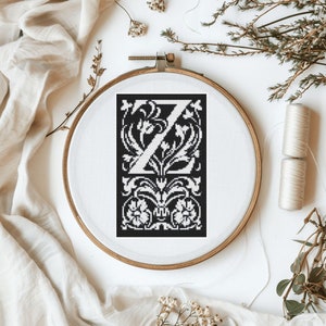 Könnte beinhalten: Ein schwarzer und weißer Stickrahmen mit einem Kreuzstich-Design des Buchstabens "Z", umgeben von verzierten Blumenmustern.