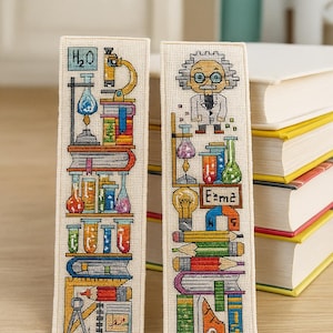 以下が含まれることがあります： 科学をテーマにした刺繍のブックマークが2つ。1つは顕微鏡、ビーカー、化学式H2Oが描かれています。もう1つは科学者、ビーカー、方程式E=mc2が描かれています。背景には本があります。