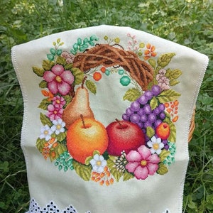 May include: A white embroidered kitchen towel with a wreath of fruit and flowers. The wreath includes a pear, apples, grapes, and pink flowers. The towel has a white crocheted border.