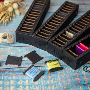 Puede incluir: Cuatro organizadores de hilo de madera negra con divisores dorados, llenos de carretes de hilo de colores. Los organizadores están sobre una superficie de madera azul, junto con carretes de hilo de varios colores, cinta de encaje y un dedal. Un tema de suministros de artesanía.