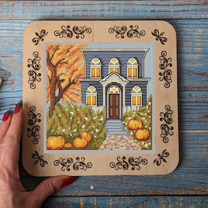 May include: A cross-stitch pattern of a blue house with white trim and a porch. The house is surrounded by a green bush and orange pumpkins. The pattern is framed in a wooden frame with decorative scrollwork.