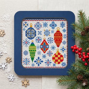 May include: A framed cross-stitch featuring festive Christmas ornaments and snowflakes. The ornaments are in various colours, including blue, red, green, and yellow. The frame is a square shape and is painted blue. The design is on a white background.