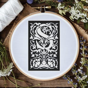Könnte beinhalten: Schwarzweißes Kreuzstichmuster eines stilisierten Buchstabens 'S' mit aufwendigen floralen Designs. Das Muster ist in einem weißen Stickrahmen gerahmt.