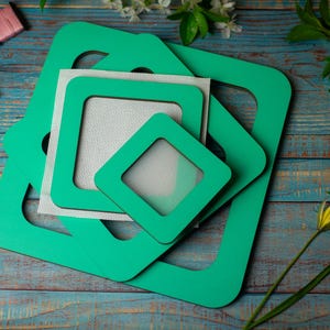Può includere: Una collezione di cornici quadrate verdi per punto croce di varie dimensioni, impilate su una superficie di legno blu invecchiato. Diversi rocchetti di filo colorato sono sparsi intorno alle cornici, insieme a rametti di fiori bianchi.