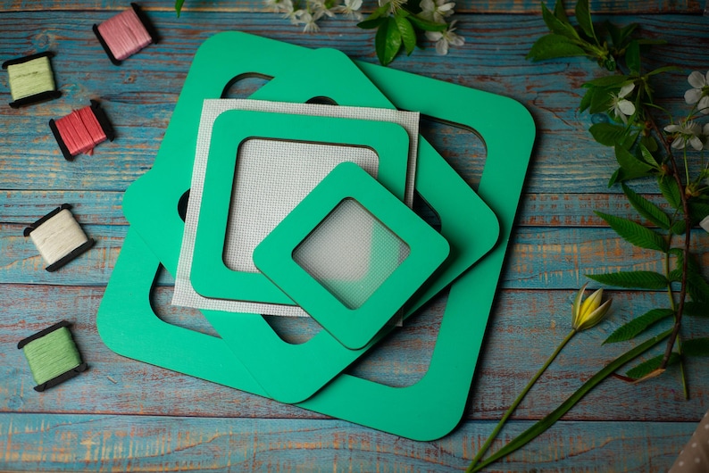 Op de afbeelding: Een verzameling groene vierkante kruissteekframes in verschillende maten, gestapeld op een verweerd blauw houten oppervlak. Verschillende spoelen kleurrijk garen liggen verspreid rond de frames, samen met takjes witte bloemen.