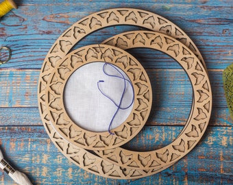 May include: Two wooden embroidery hoops with intricate floral designs. The smaller hoop is inside the larger hoop and both are on a blue wooden surface.