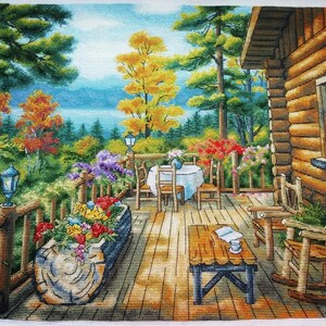 May include: A cross-stitch landscape depicts a cabin porch with a table set for dining, surrounded by colorful flowers and trees. A wooden rocking chair and a small table with a book and a cup are also visible. The scene is set in a forest with a lake in the background.