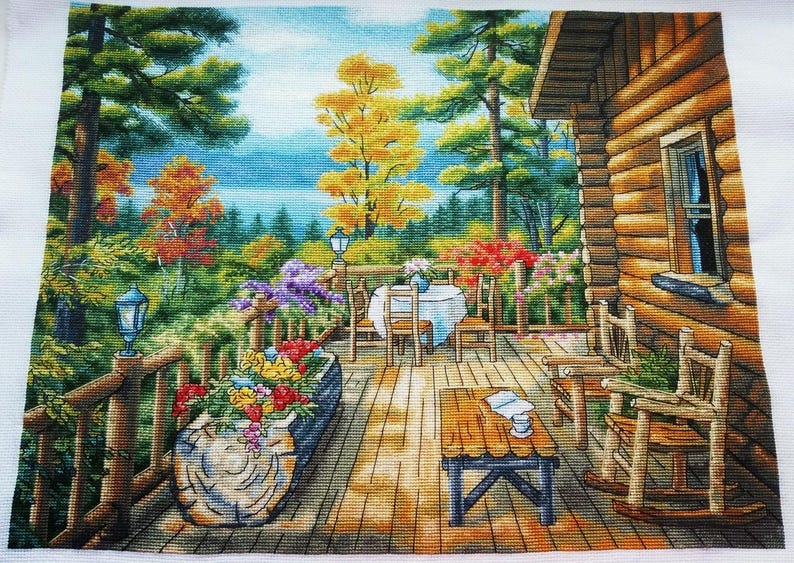 May include: A cross-stitch landscape depicts a cabin porch with a table set for dining, surrounded by colorful flowers and trees. A wooden rocking chair and a small table with a book and a cup are also visible. The scene is set in a forest with a lake in the background.