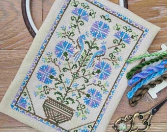 Puede incluir: Un patrón de punto de cruz que presenta un diseño floral estilizado con flores azules, moradas y verdes en una maceta marrón. El patrón está enmarcado por un borde marrón.