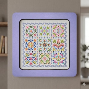 May include: A framed cross-stitch sampler with a lavender-colored wooden frame. The sampler features nine colorful floral designs in a grid pattern, with a decorative border. The embroidery uses a variety of colors including pink, blue, yellow, and green.