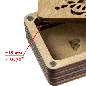 May include: Close-up of a wooden box with a hinged lid. The box is made of layered wood with a dark brown stain. The lid has a decorative cutout design. The box is approximately 0.71 inches deep.