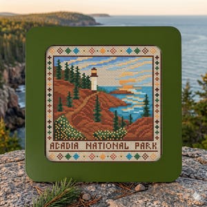 May include: A square cross-stitch embroidery in a dark green frame depicting a coastal landscape with a lighthouse, evergreen trees, and a sunset over the water. The text ACADIA NATIONAL PARK is stitched at the bottom with colorful geometric borders.