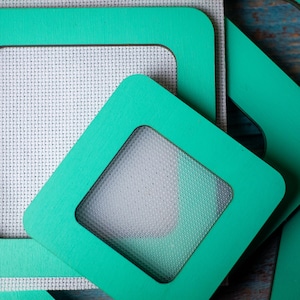 Op de afbeelding: Close-up van verschillende turquoise vierkante frames met afgeronde hoeken. De frames hebben een vensteruitsparing, die een witte, getextureerde stof onthult. De frames zijn gerangschikt in een gelaagde compositie.