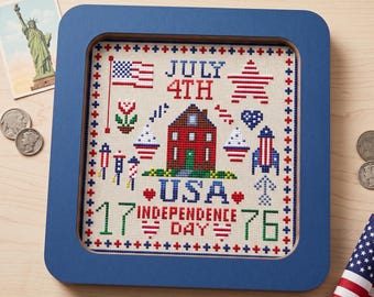 May include: A framed cross-stitch featuring patriotic symbols for the 4th of July. The design includes a red house, American flag, fireworks, and the text "USA 1776 Independence Day". The frame is a square shape with a blue finish.