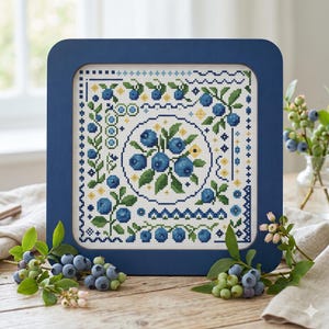 May include: A framed cross-stitch featuring a blueberry design. The artwork is set in a square, navy blue frame. The cross-stitch showcases a central blueberry cluster surrounded by green leaves and yellow accents, with a white background.