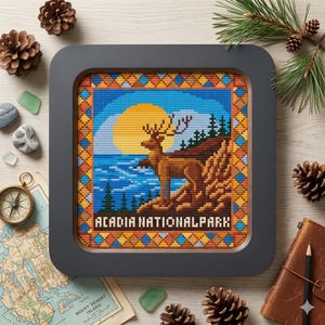 May include: A framed cross-stitch depicting a deer overlooking a body of water with a large sun in the background. The text "ACADIA NATIONAL PARK" is stitched at the bottom. The frame is dark gray.