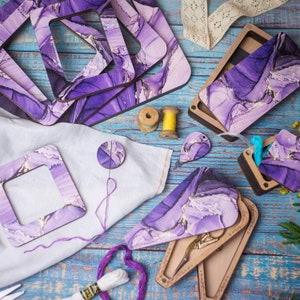 May include: An assortment of craft supplies, including wooden frames, a needle minder, and scissor cases, all featuring a purple and gold marbled design. The items are arranged on a blue wooden surface, with spools of thread and embroidery floss.