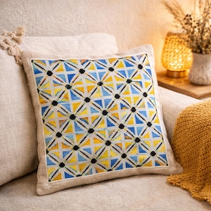 May include: Square decorative pillow with a geometric pattern in yellow, blue, and black. The pillow has a beige border and is placed on a cream-colored sofa. The pillow's design features triangles and dots.