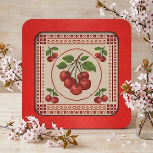 May include: A framed cross-stitch featuring a cluster of red cherries with green leaves, set against a cream background. The artwork is enclosed in a square, red wooden frame, with cherry blossom branches in the background.