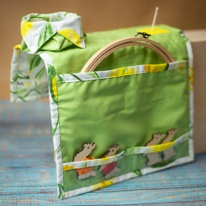May include: Green fabric organizer with a pocket featuring wooden bunny cutouts. The organizer has a wooden embroidery hoop and is trimmed with white and yellow floral fabric. The background is a wooden box.
