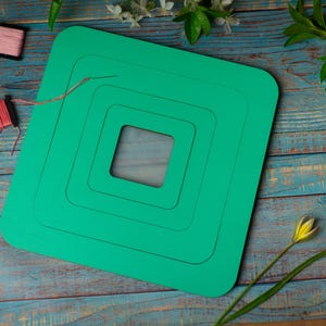 Op de afbeelding: Een turquoise vierkante knutselmat met een genest vierkant ontwerp, omringd door spoelen garen in verschillende kleuren. De mat ligt op een blauw houten oppervlak, met bloemaccenten.