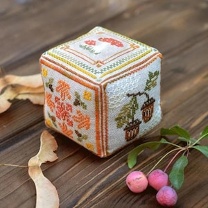 Könnte beinhalten: Ein weißer Stoffwürfel mit einem Kreuzstich-Design mit Herbstblättern, Eicheln und Pilzen. Der Würfel liegt auf einer Holzoberfläche mit einem Zweig kleiner roter Äpfel.